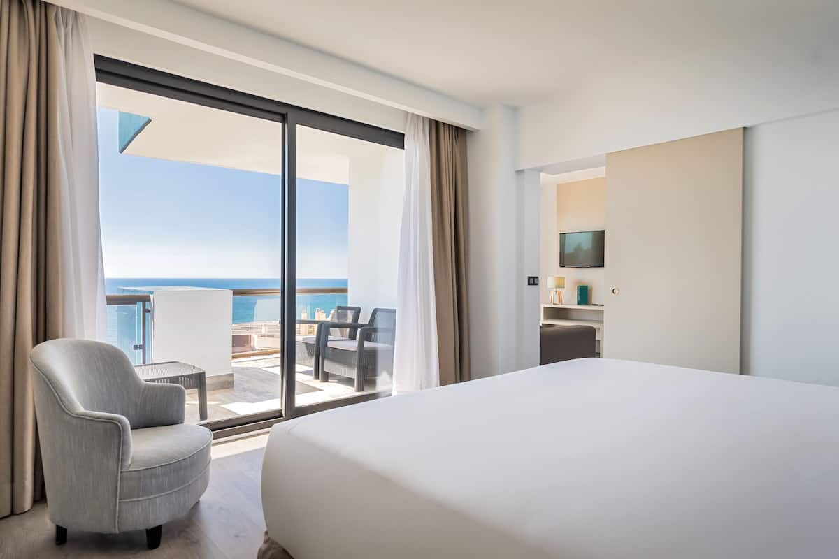A hotel room with a large bed, a chair, a TV, and a balcony with a view of the sea.