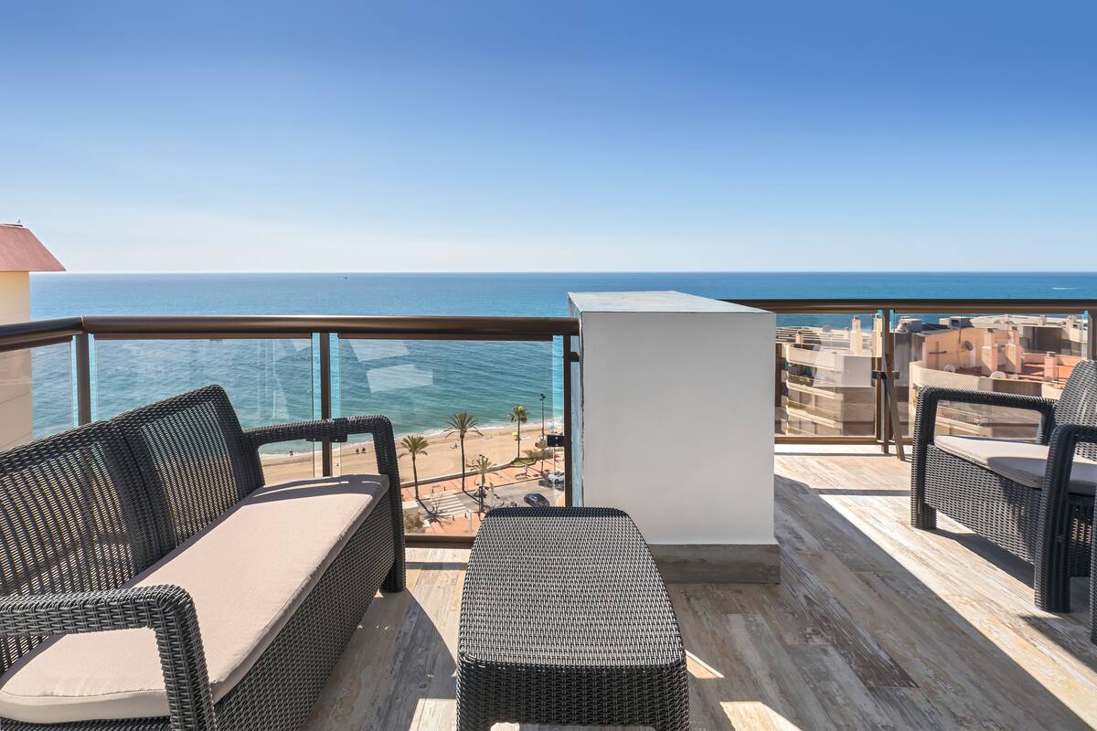 A balcony with wicker furniture, a view of the sea, and a cityscape in the background.