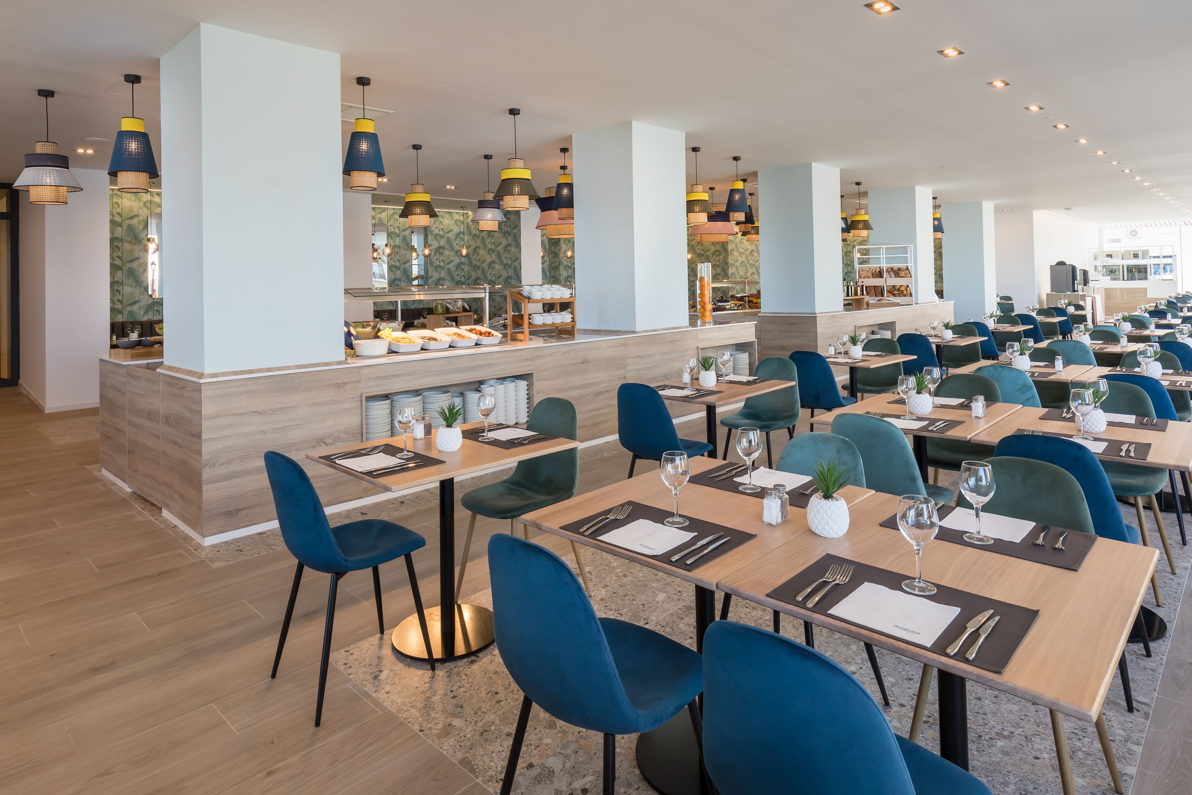 Ein geräumiges Restaurant mit mehreren Tischen und Stühlen, einer zentralen Theke mit Speisenauslage und moderner Pendelleuchten.