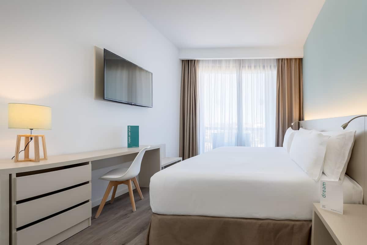 A hotel room with a bed, a desk, a chair, a TV, and a window with curtains.