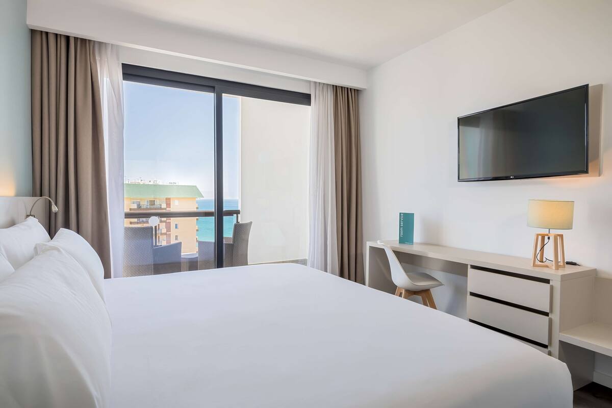 A hotel room with a large bed, a desk, a chair, a TV, and a view of buildings through the window.