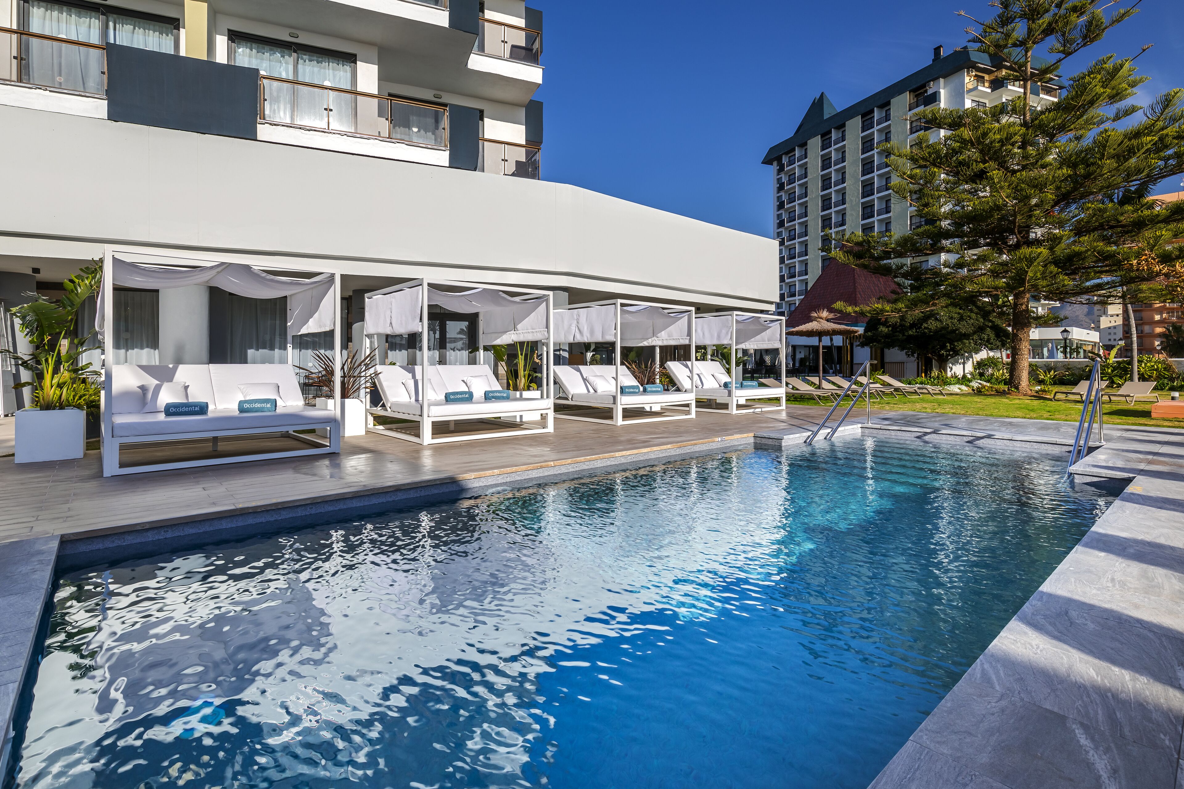 Un espace aménagé au bord de la piscine, avec des chaises longues et des parasols, une piscine à l’eau d’un bleu limpide et un bâtiment moderne en arrière-plan.