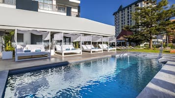 Un área junto a la piscina con sillones de descanso y sombrillas, una piscina azul cristalina y un edificio moderno al fondo.
