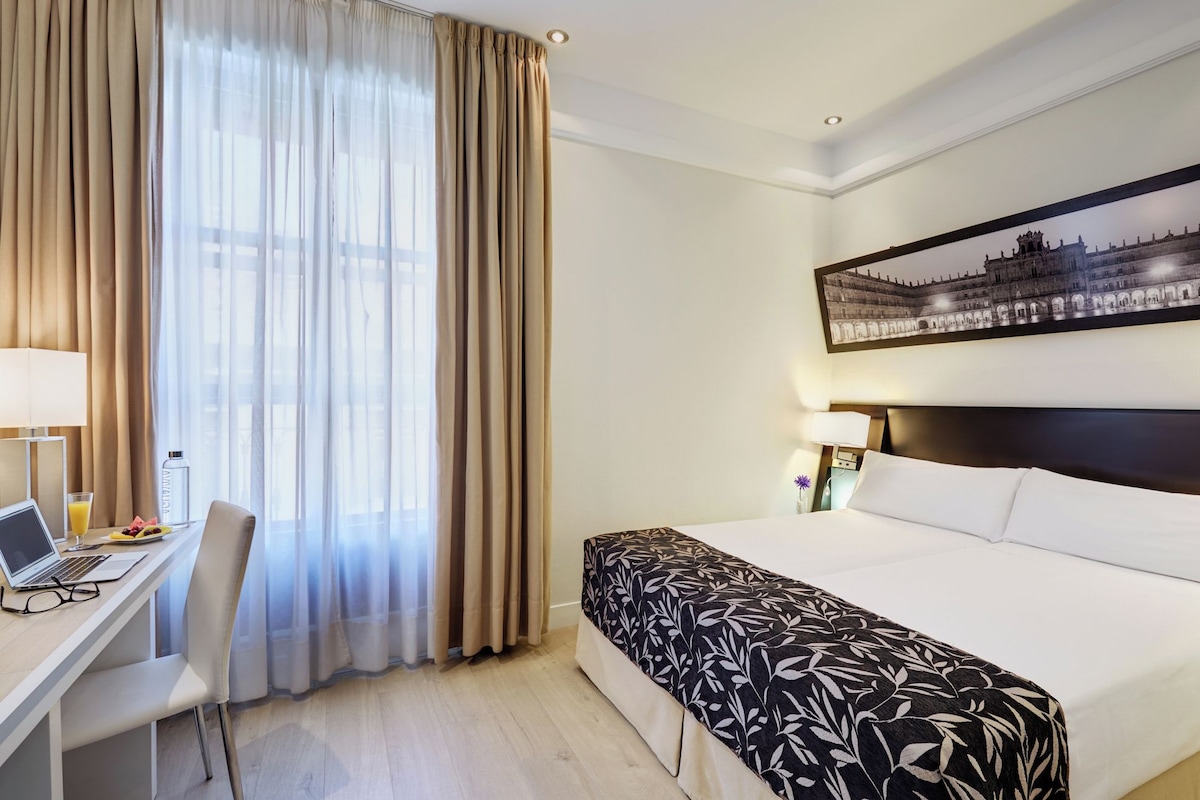 Ein Hotelzimmer mit Bett, Schreibtisch mit Laptop, Stuhl, Nachttisch und einem großen Fenster mit Vorhängen.