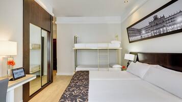 Habitación de hotel con una cama grande, una litera, un escritorio con ordenador, un espejo y un cuadro en la pared.
