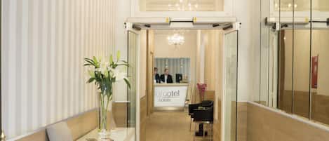 A hotel reception area with a reception desk, a chandelier, a seating area with a vase of flowers, and a mirror wall.