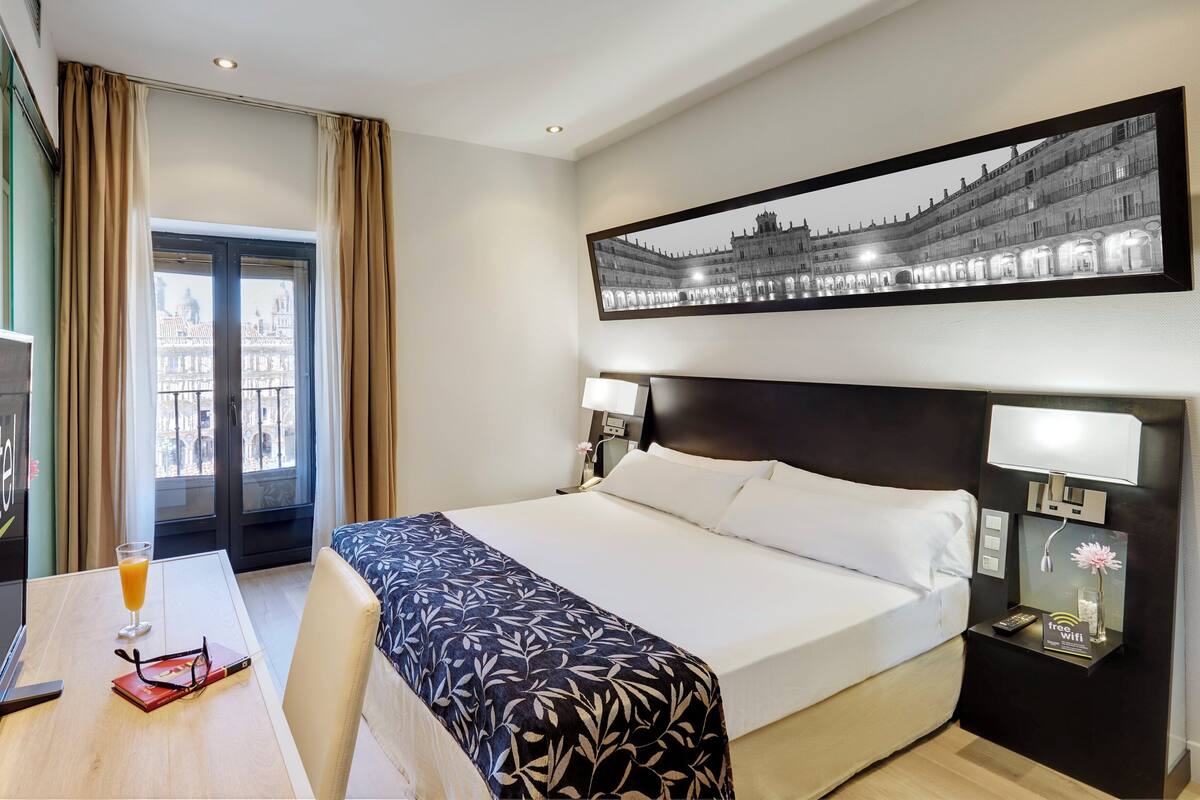 A hotel room with a bed, bedside lamps, a desk with a glass of orange juice, a book, and a framed picture on the wall.