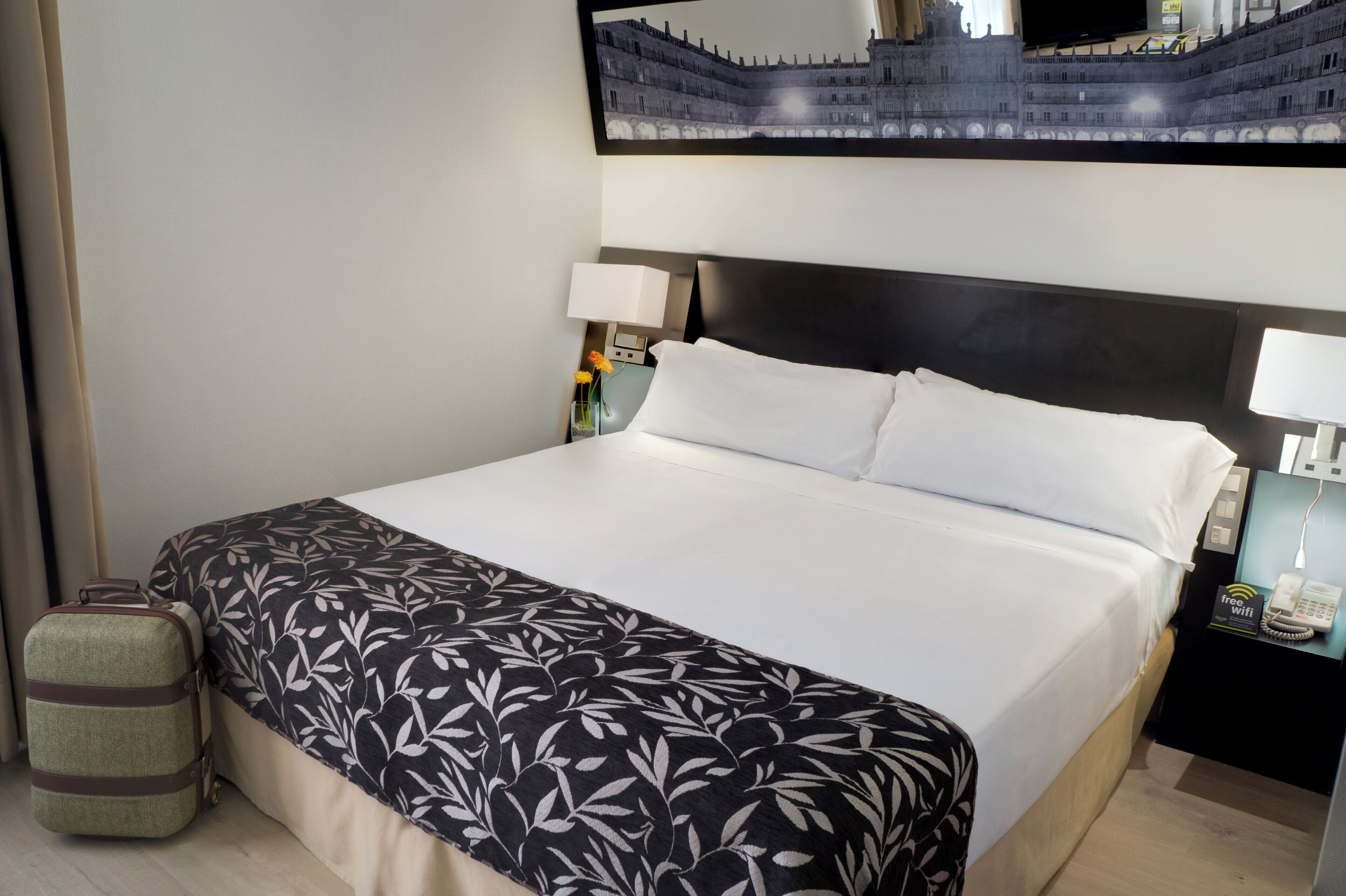 Une chambre d’hôtel avec un grand lit, une table de chevet avec une lampe, une petite table sur laquelle est posé un vase de fleurs et un téléviseur fixé au mur.
