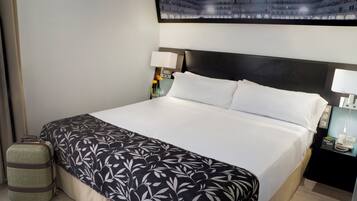 Una habitación de hotel con una cama grande, una mesita de noche con una lámpara, una pequeña mesa con un jarrón de flores y un televisor montado en la pared.