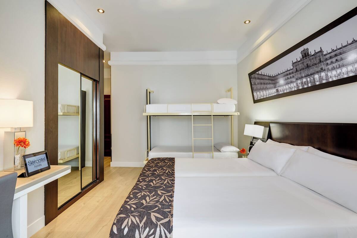 A hotel room with a large bed, a bunk bed, a desk with a computer, a mirror, and a framed picture on the wall.