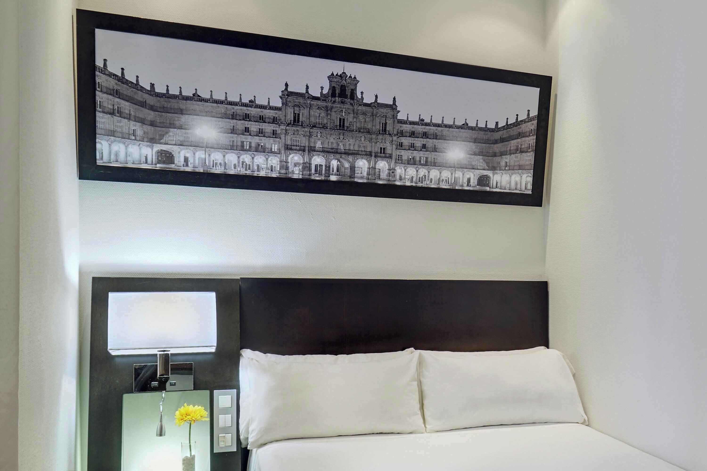 A hotel room with a large framed picture of a historic building, a bed with white linens, a nightstand with a lamp and a vase of flowers.