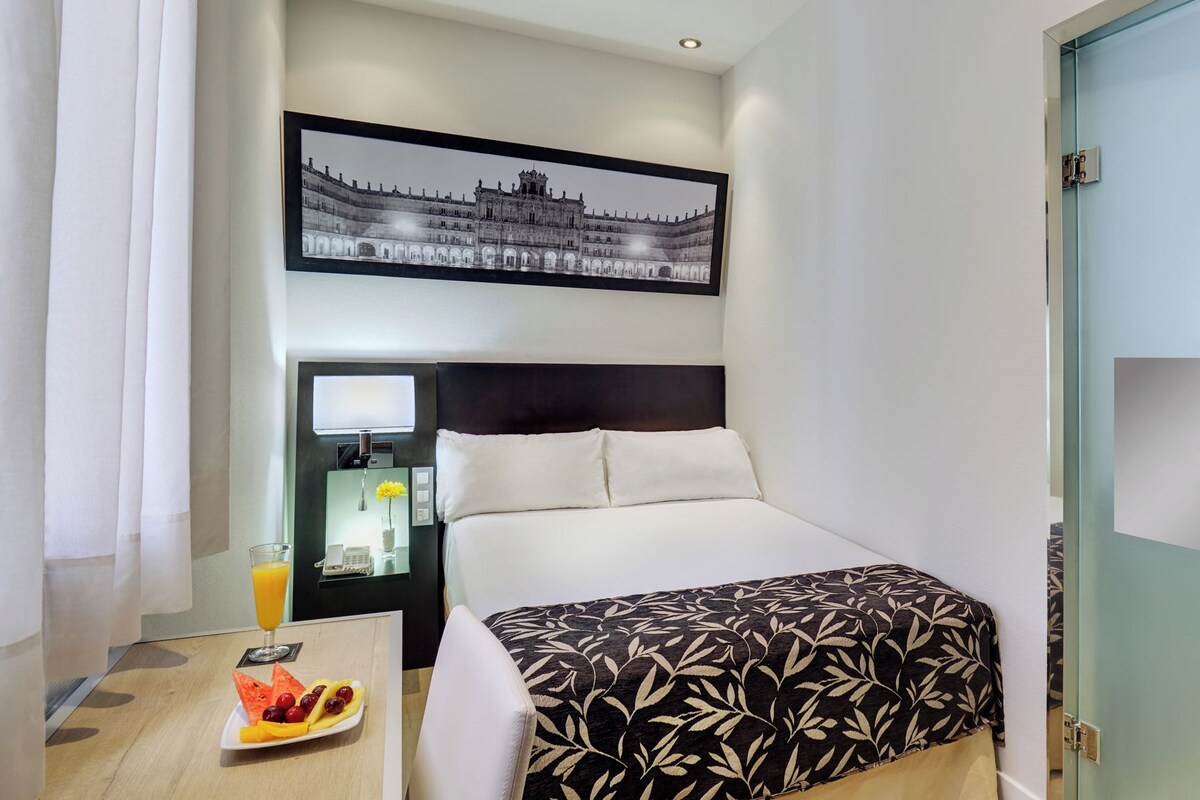 Une chambre d’hôtel avec un lit, une table de chevet avec une lampe, un tableau encadré au mur et un plateau de petit-déjeuner sur la table.
