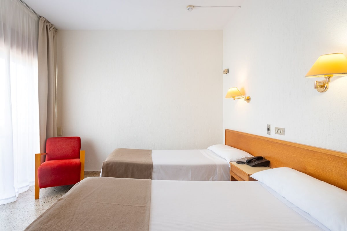 Une chambre d’hôtel avec deux lits, un fauteuil rouge et deux lampes fixées au mur.