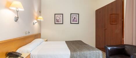 A hotel room with a bed, bedside table with a phone, two wall-mounted lamps, and two framed pictures.