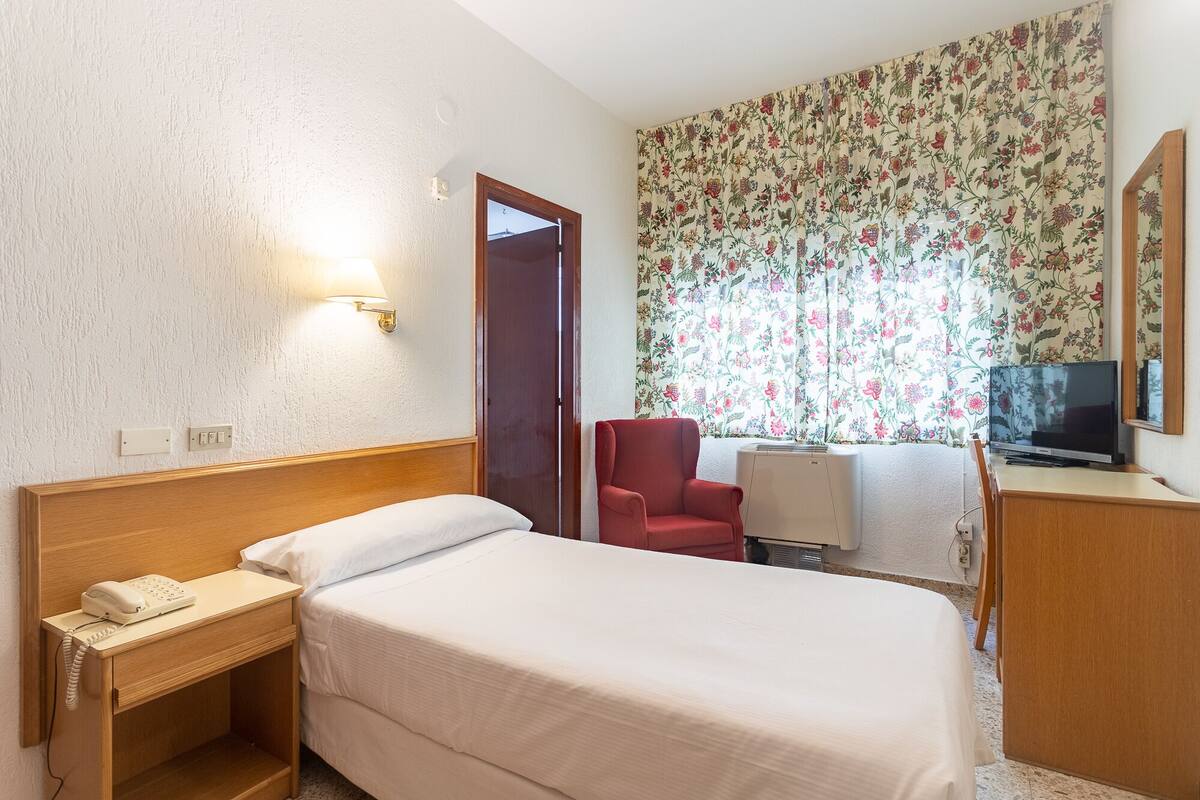 Une chambre d’hôtel comprenant un lit, un bureau avec un téléphone, une chaise, un téléviseur et une fenêtre avec des rideaux à motifs floraux.