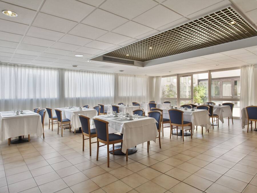 Un comedor amplio con varias mesas y sillas, ventanas grandes con cortinas y un techo con diseño de cuadrícula.