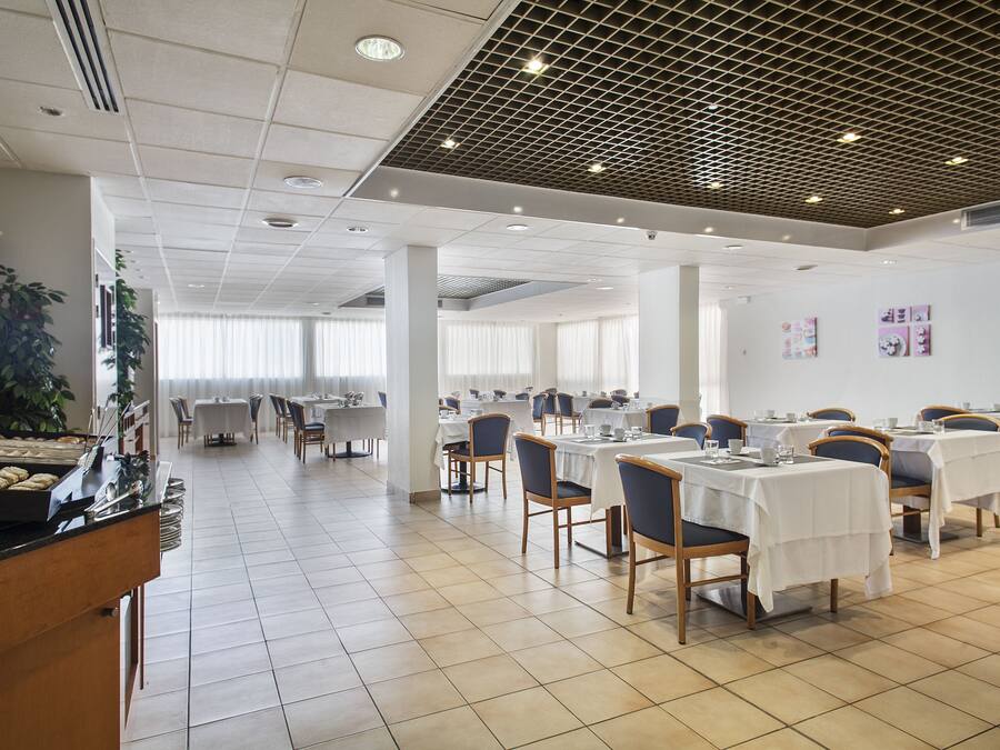A spacious dining area with multiple tables and chairs, a kitchen counter with baked goods, and large windows with curtains.