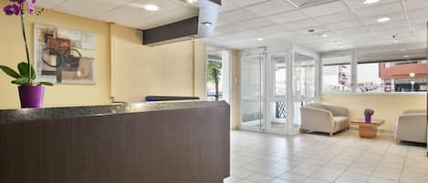 A reception area with a dark reception desk, a purple orchid, and a painting on the wall. There are two chairs and a small table with a plant.