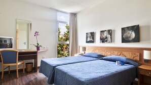 Une chambre d’hôtel avec deux lits, un bureau, une chaise, une plante en pot et des œuvres d’art accrochées au mur.