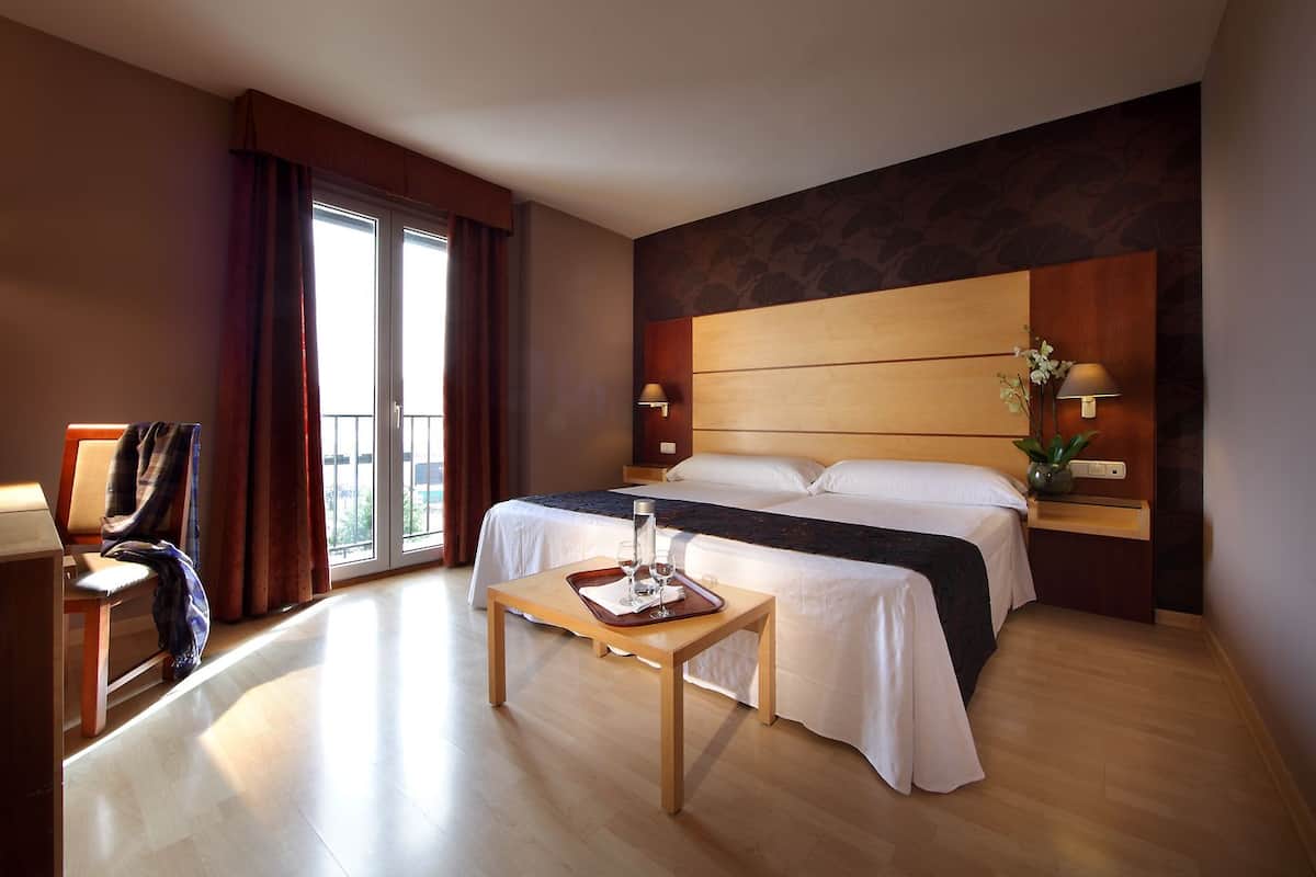 Une chambre d’hôtel avec un grand lit, une petite table avec un verre et un plateau, une chaise, un bureau et une fenêtre avec des rideaux.