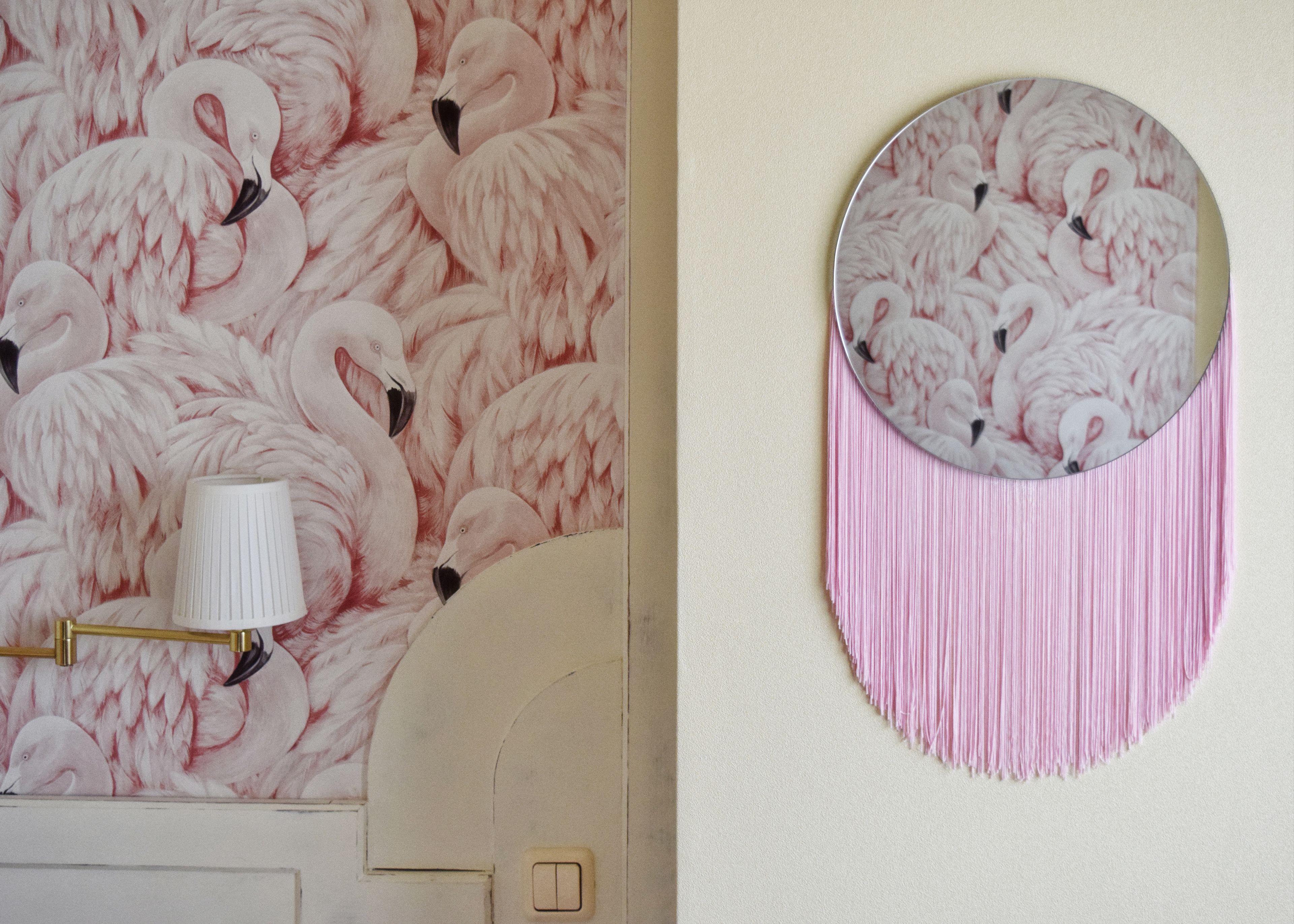 A wall with a flamingo wallpaper and a round mirror with pink fringes.