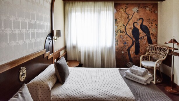 A hotel room with a bed, a chair, a mirror, a wall with a tree and birds mural, and a window with curtains. - Hotel María Luisa (Burgos)