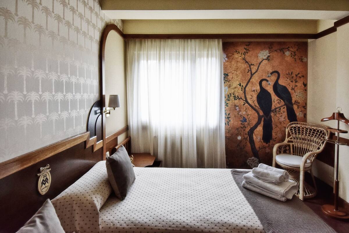 A hotel room with a bed, a chair, a mirror, a wall with a tree and birds mural, and a window with curtains.