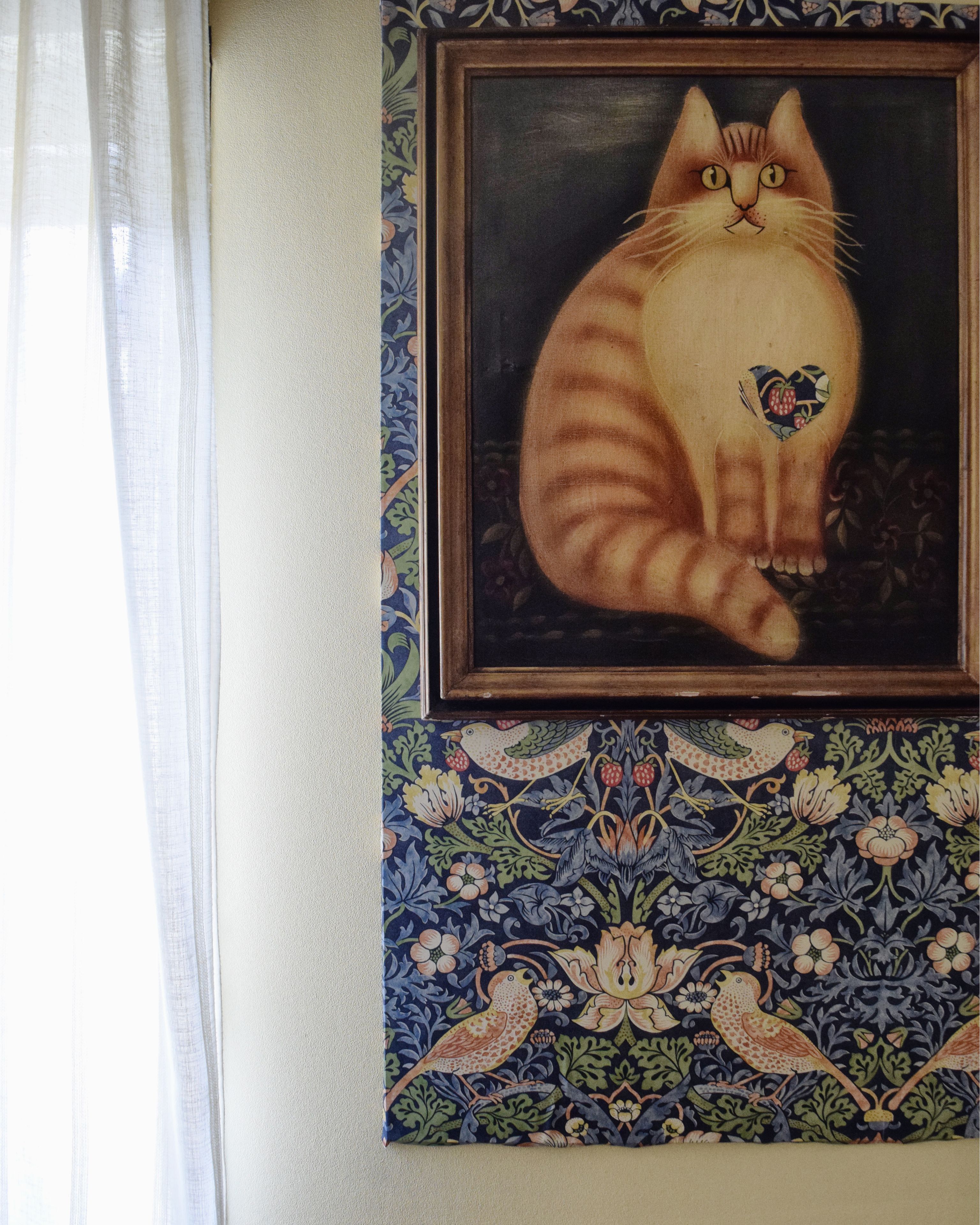 A framed painting of a cat with a heart on its chest, set against a wall with a floral patterned border.
