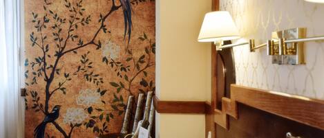 Ein Hotelzimmer mit einem Bett, einem Nachttisch, einer Lampe, einem an der Wand befestigten Regal und einer Wand mit Vogel- und Blumendekor.