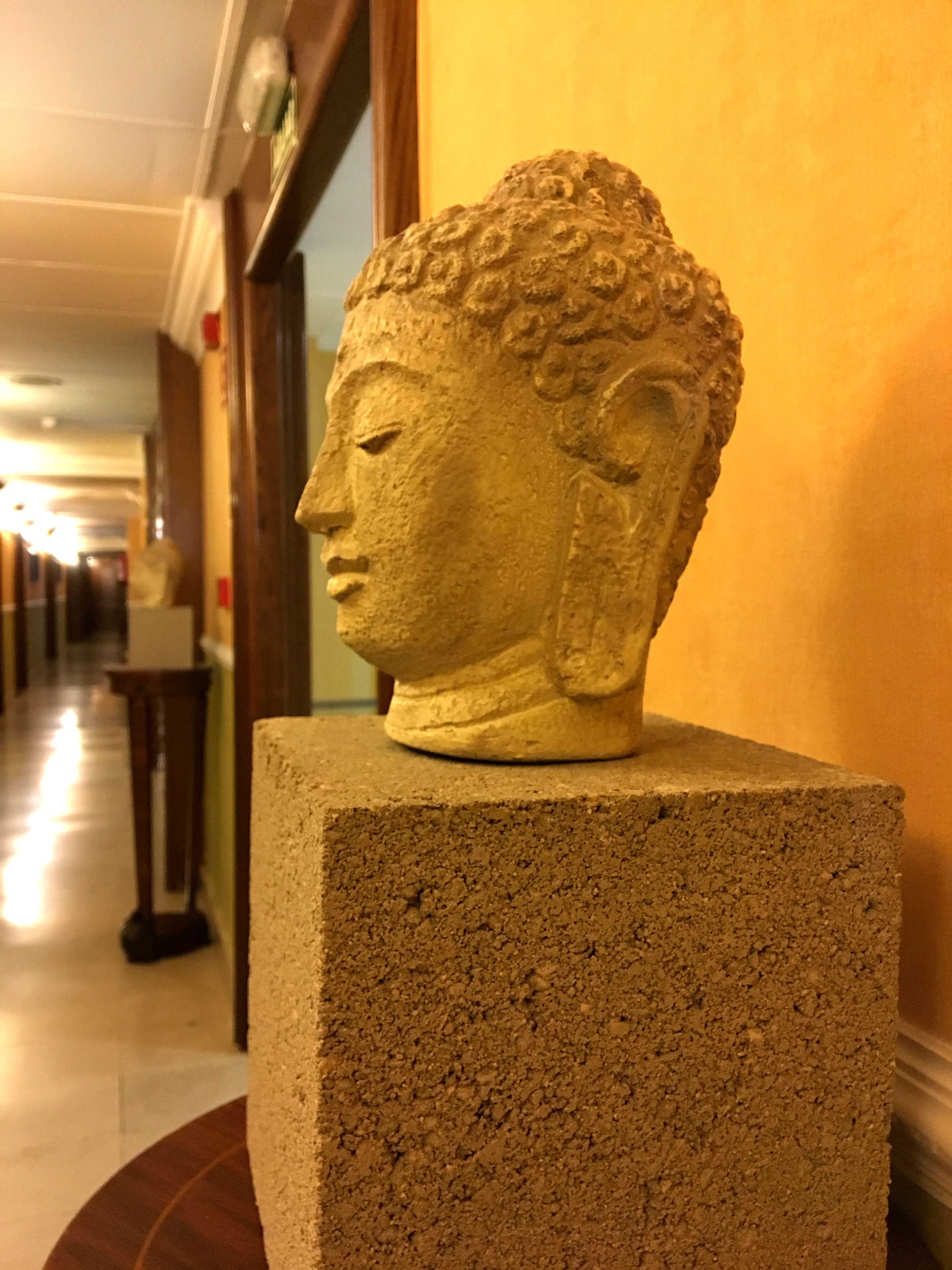 Una escultura de cabeza de Buda de piedra sobre un pedestal en un pasillo con suelo de madera y pared amarilla.