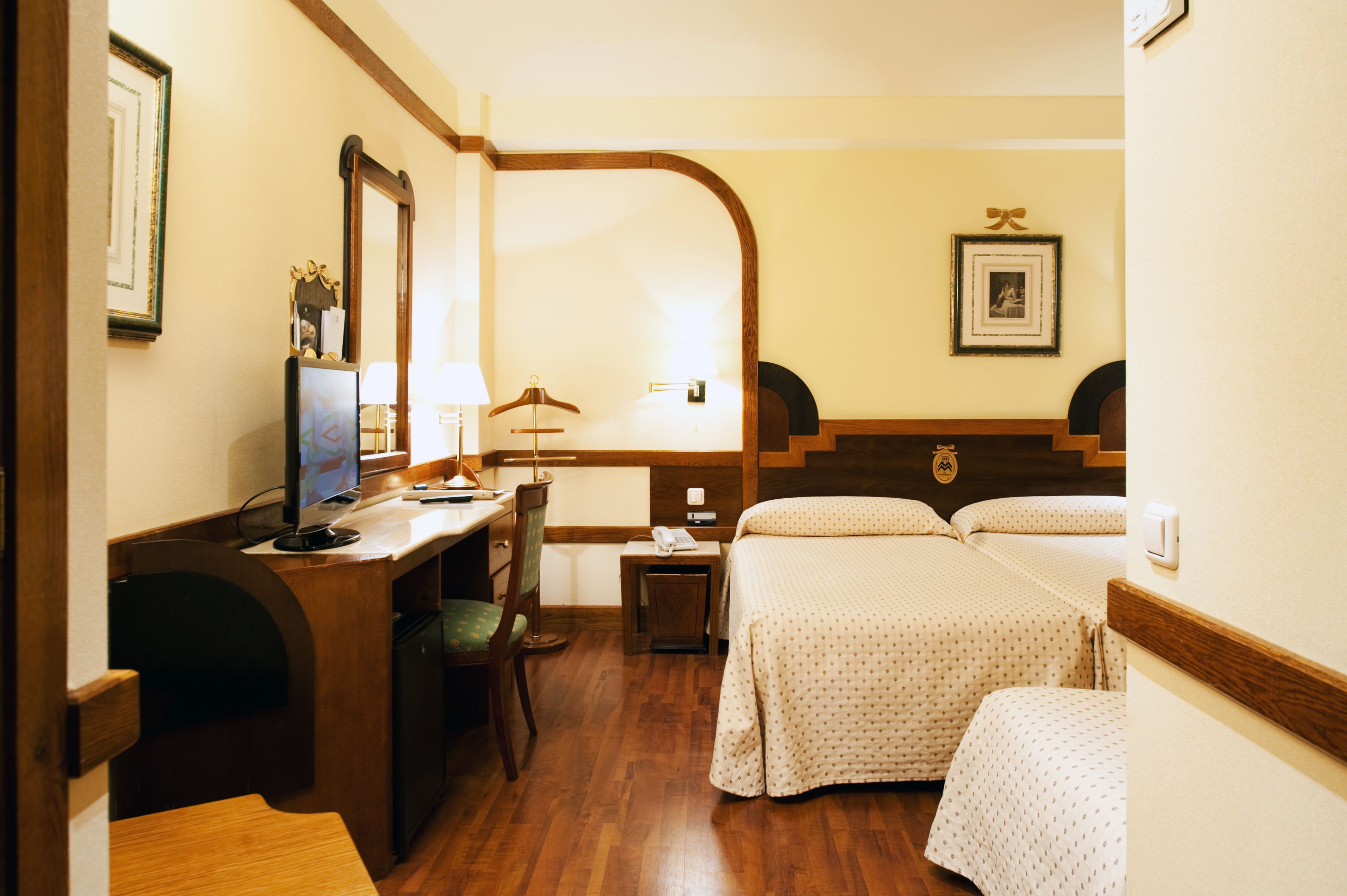A hotel room with a wooden desk, a chair, a bed with a headboard, a bedside table, and a lamp.