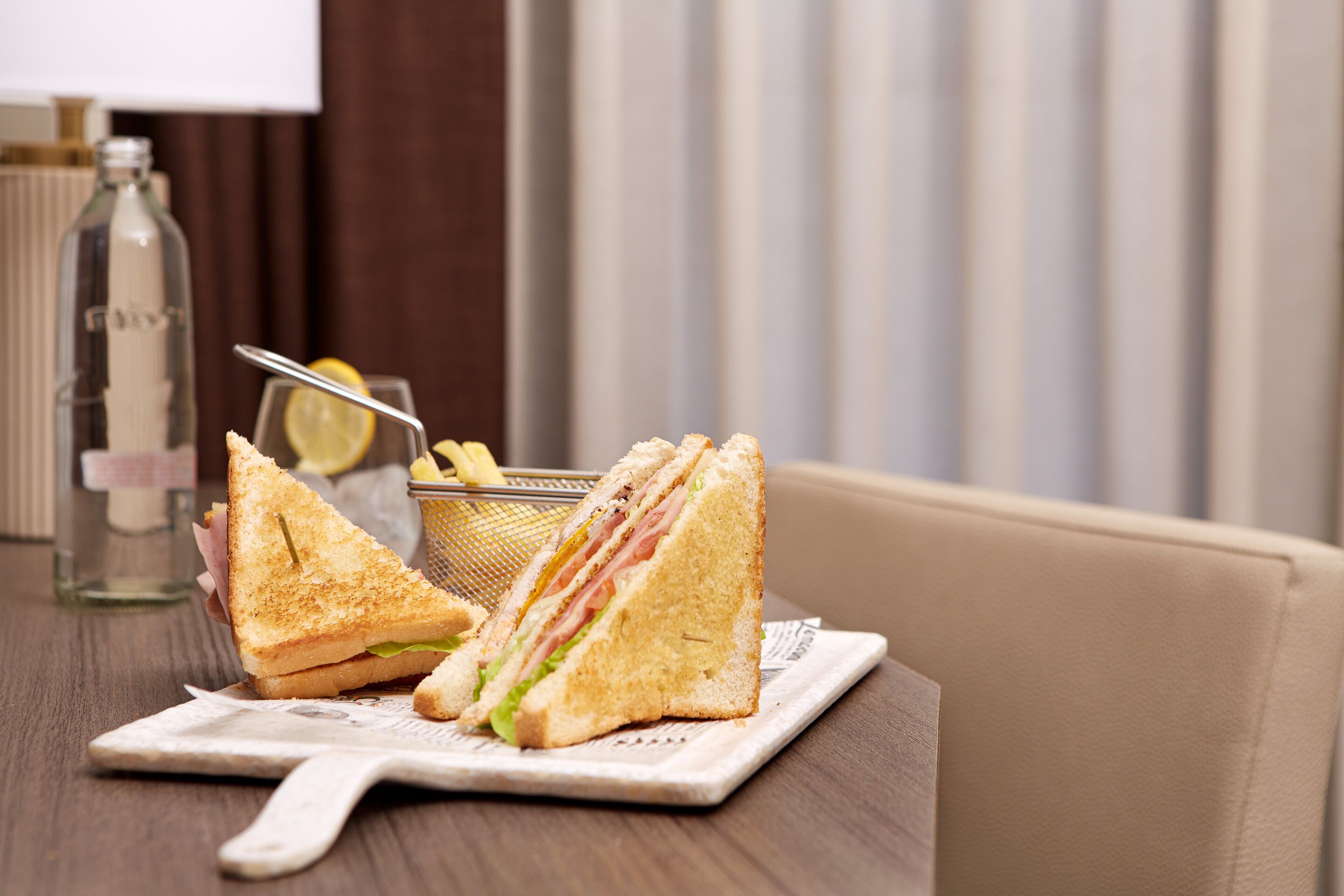 Un sándwich en un plato con un tenedor, una botella y un trozo de limón sobre una mesa.