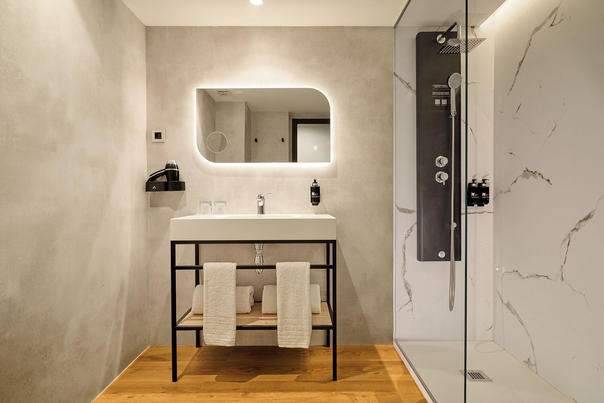 Un baño moderno con suelo de madera, un lavamanos blanco con marco negro, una ducha con mampara de vidrio y una pared de mármol.