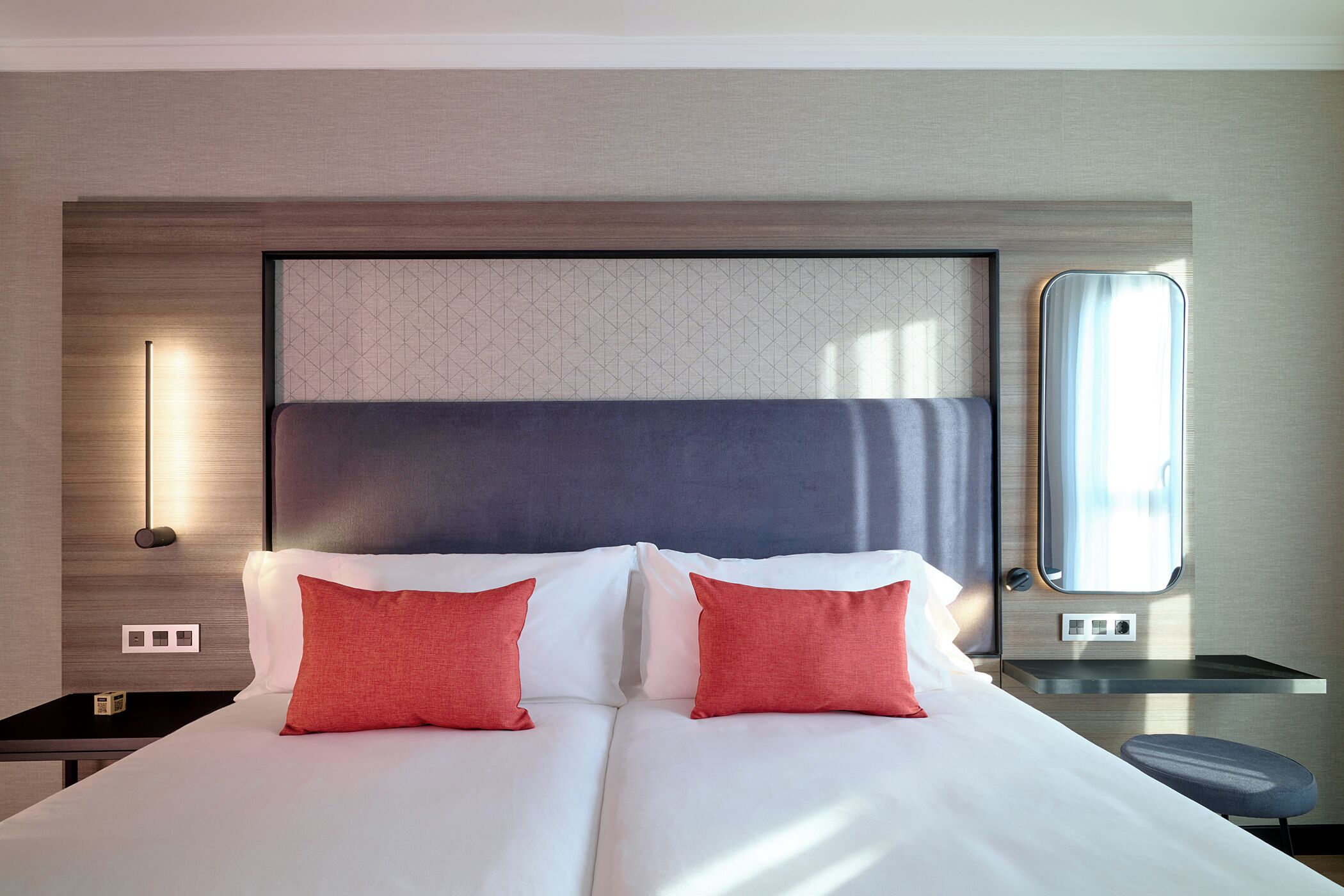 A hotel room with a large bed, two red pillows, a nightstand, a mirror, and a small table.