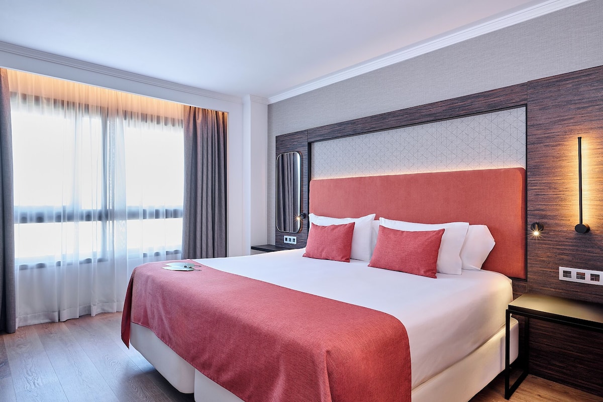 Hotelkamer met een groot bed, een nachtkastje, een wandlamp en een raam met gordijnen.