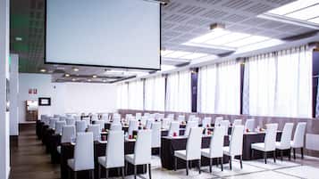 A conference room with rows of white chairs, a large projection screen, and a long table with water bottles and fruit.