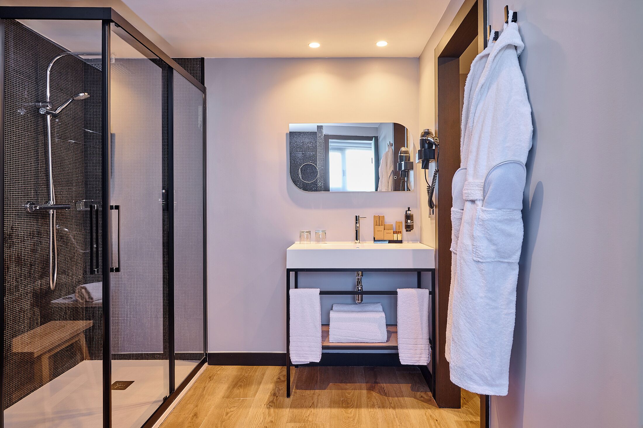 Un baño moderno con cabina de ducha de vidrio, un lavabo blanco con espejo encima y un perchero con una bata colgada.