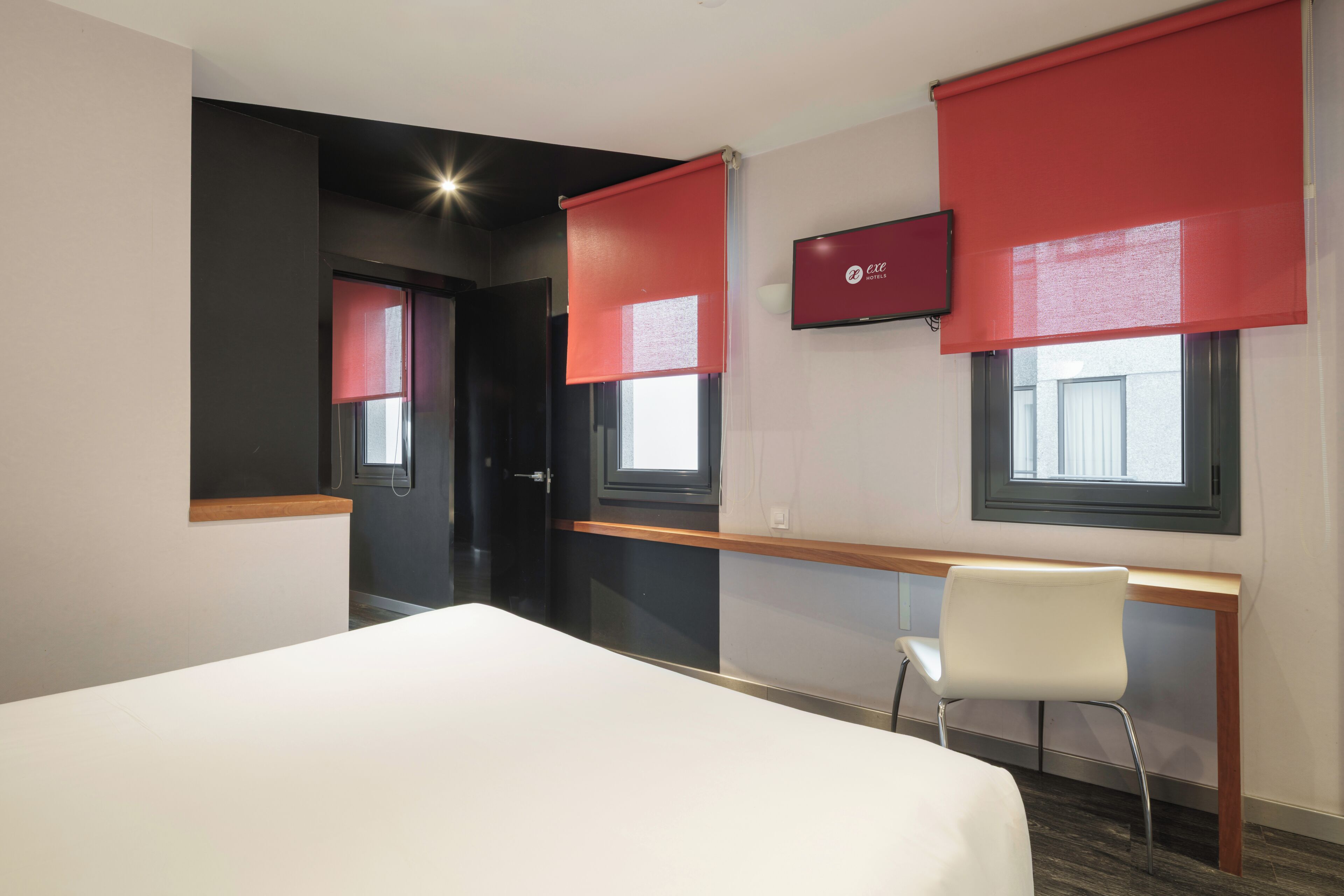 Habitación de hotel con cama, escritorio, silla, televisor y persianas rojas en las ventanas.