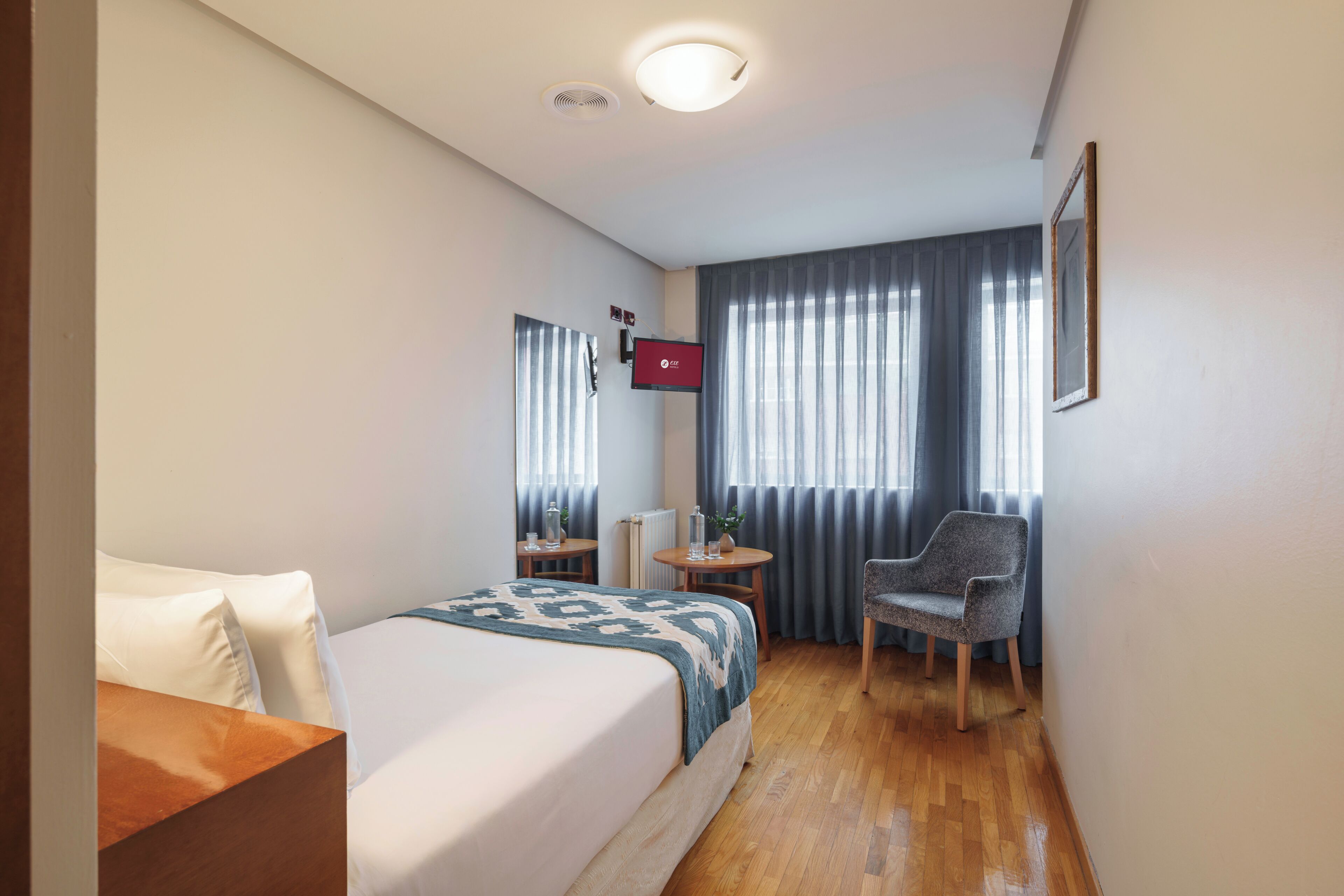 A hotel room with a bed, a desk, a chair, a TV, and a window with curtains.