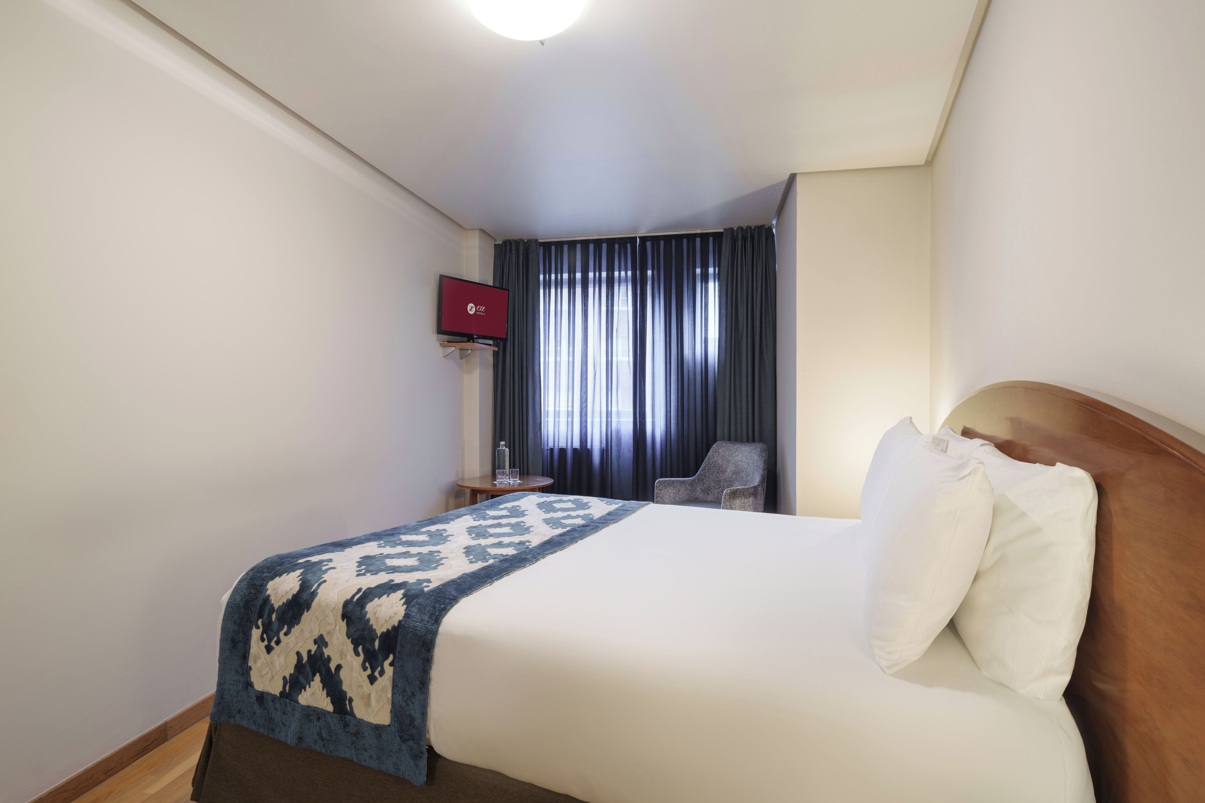 A hotel room with a bed, a TV, a chair, and a window with curtains.