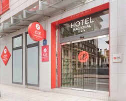 Fachada de un hotel con señalización que indica 'HOTEL MAGISTRAL' y 'exe MAGISTRAL', con un frente de vidrio y detalles en rojo.
