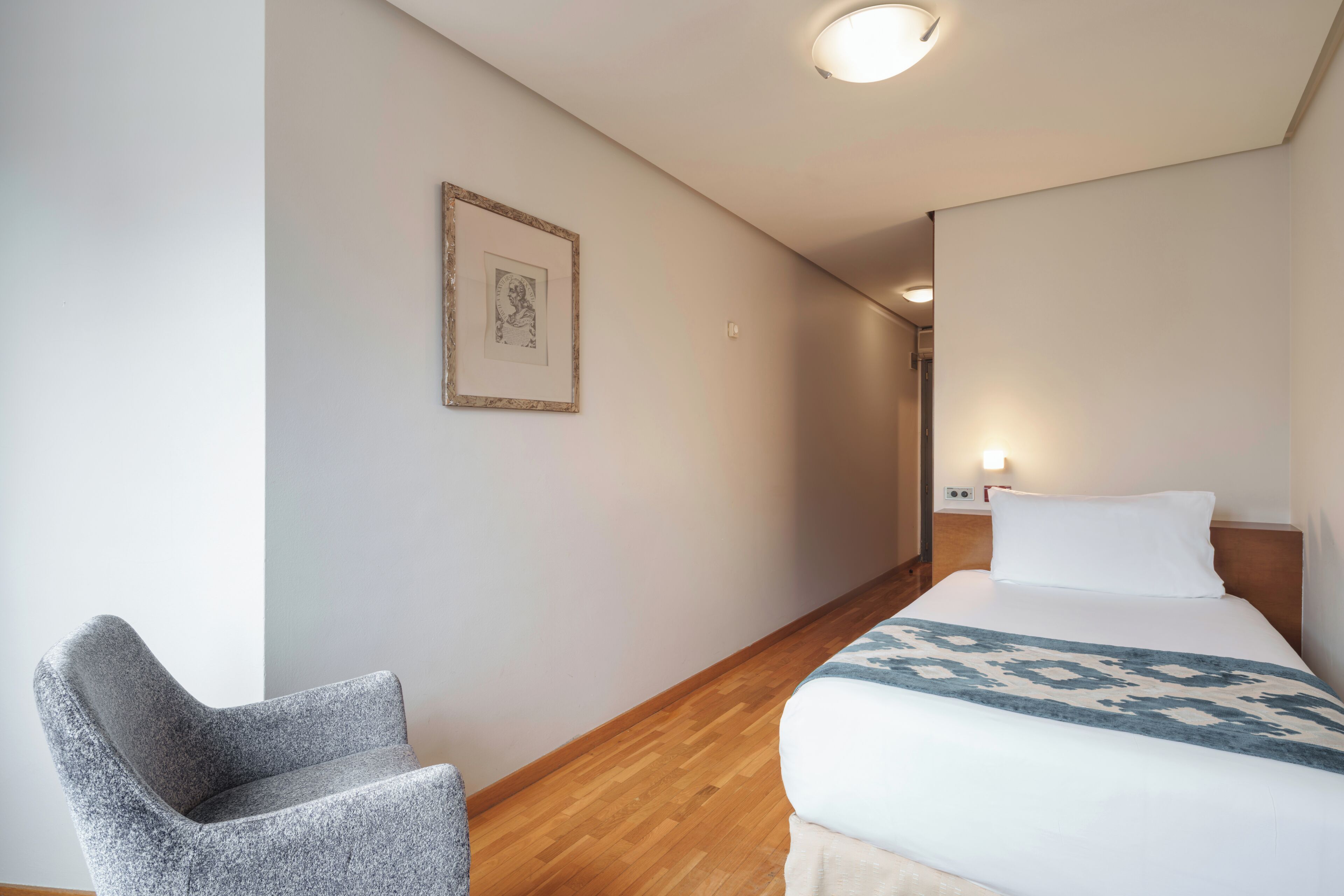Una habitación de hotel con una cama, una silla, un cuadro en la pared y un pasillo que conduce a otra habitación.