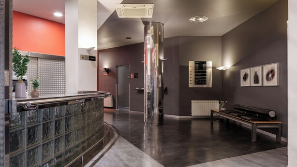 A modern lobby with a reception desk, a bench, a large pillar, and a wall-mounted information board. - Exe Hotel El Magistral (Oviedo)
