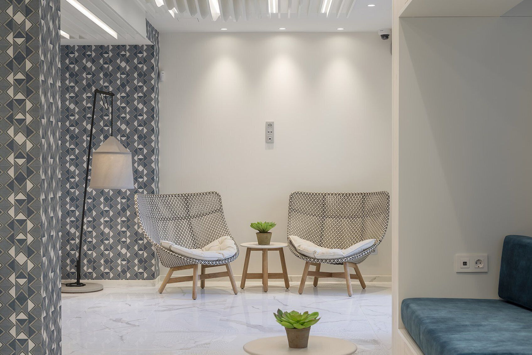 Un área de estar moderna con dos sillones de mimbre, una mesita con una planta y un sofá azul.