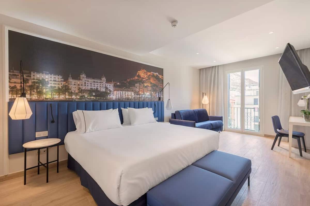 A modern hotel room with a large bed, a seating area, a desk, and a cityscape view.