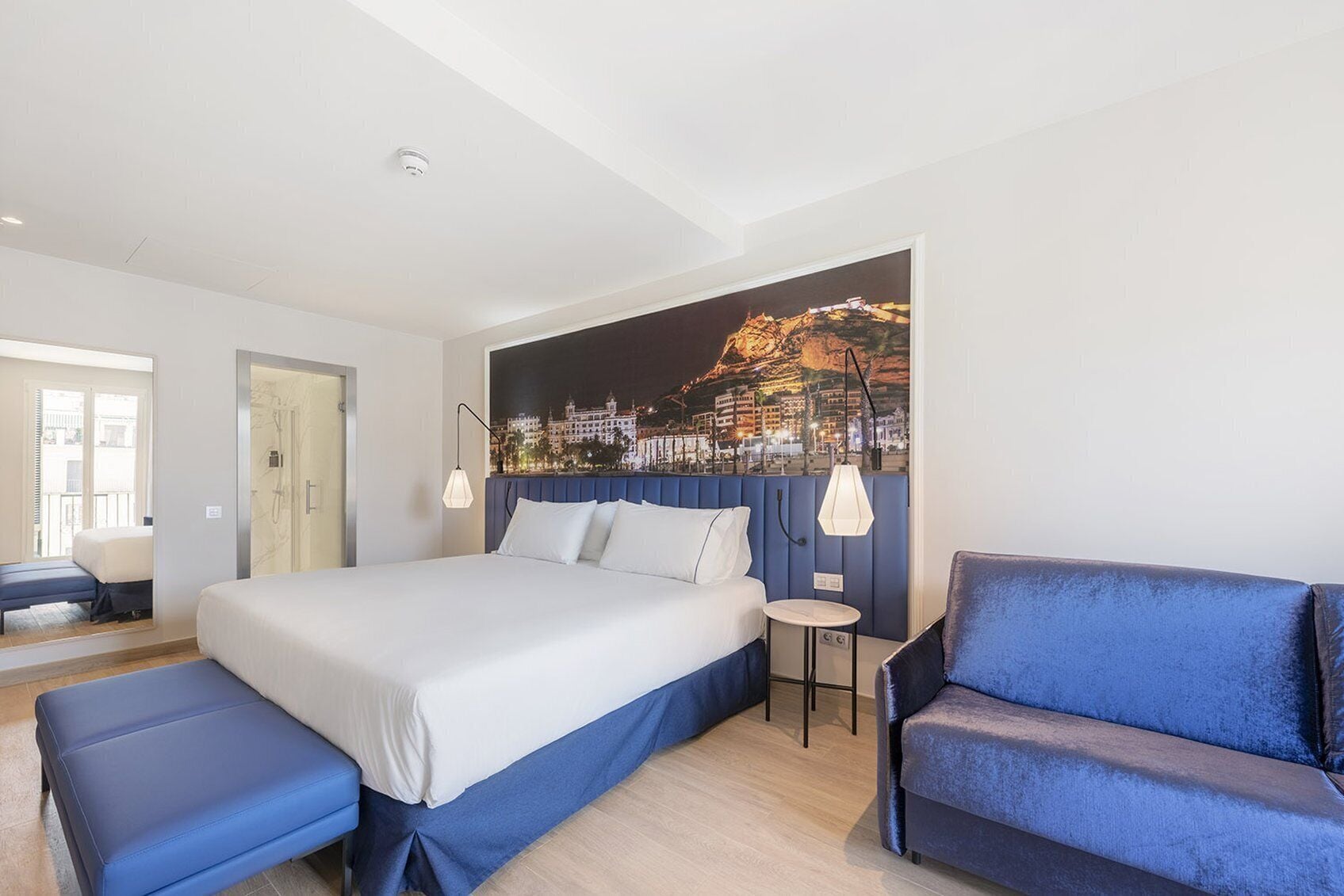 A modern hotel room with a large bed, a blue sofa, and a cityscape mural on the wall.
