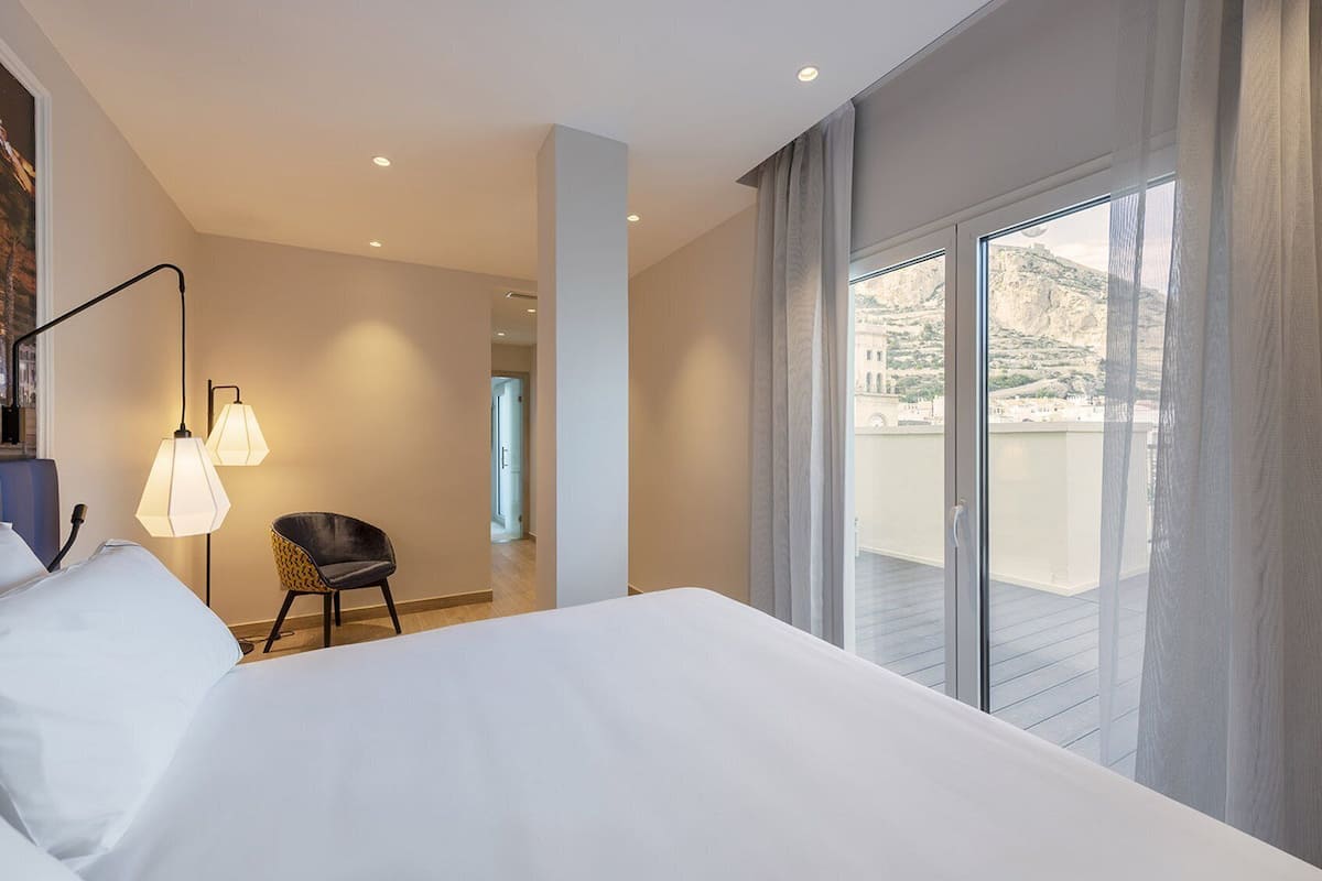 A hotel room with a large bed, a chair, a desk, and a balcony with a view.