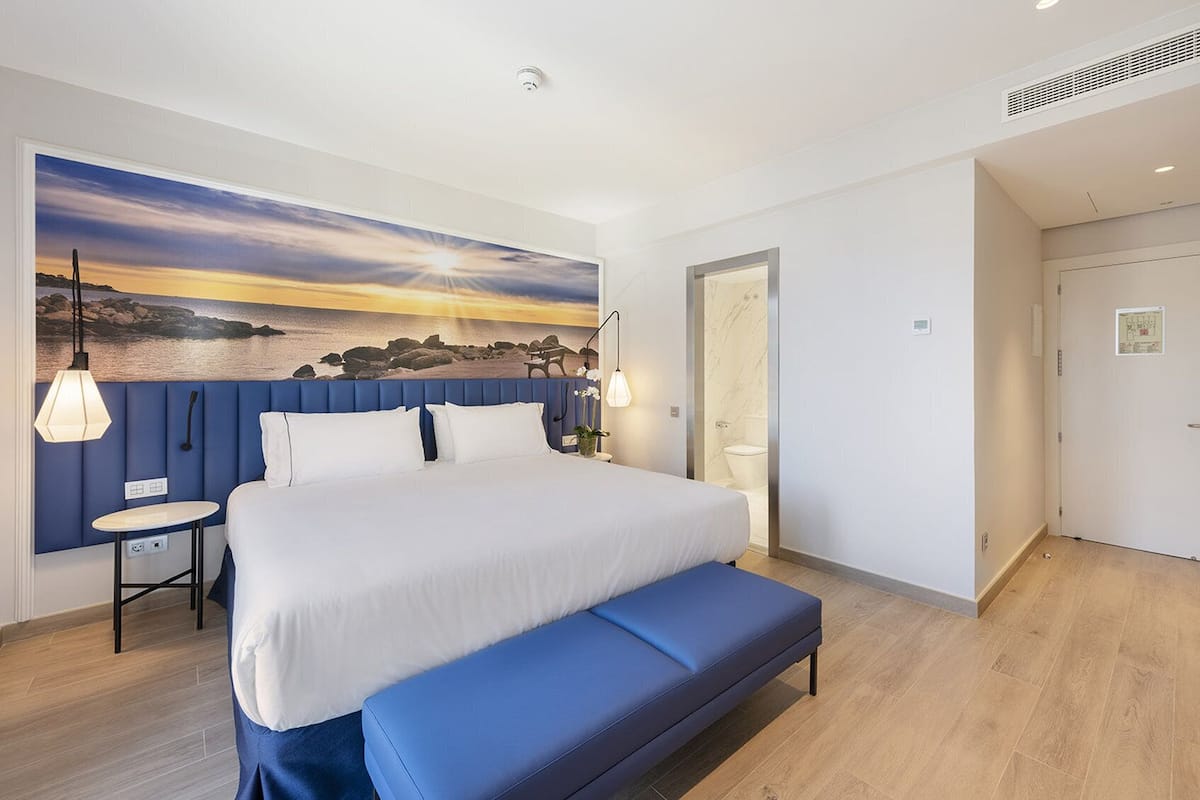 A modern hotel room with a large bed, a bench, a small table, and a painting of a sunset over the sea.