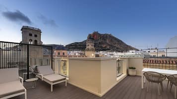 Terraza en la azotea con sillones de descanso y una mesa, con vistas a un paisaje urbano con edificios históricos y una colina destacada.