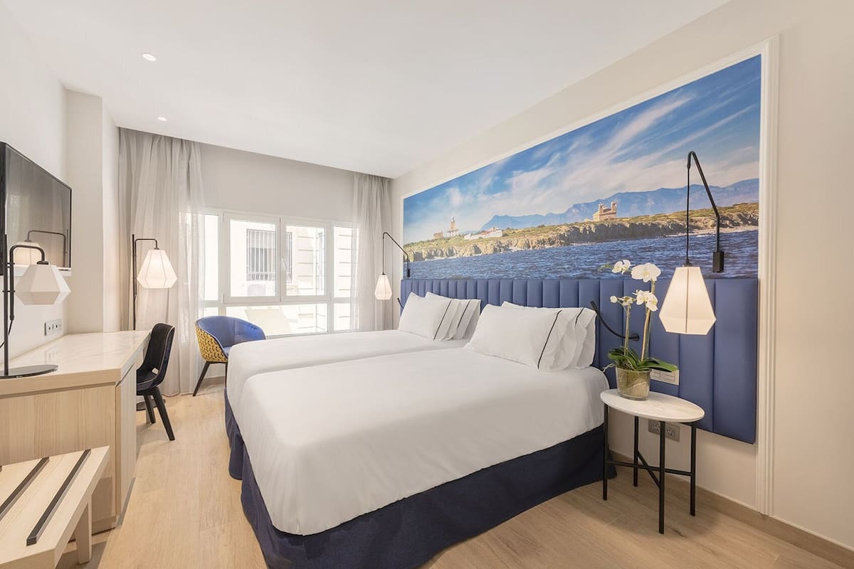 A hotel room with a large bed, a desk, a chair, a small table with a plant, and a painting of a coastal landscape on the wall.